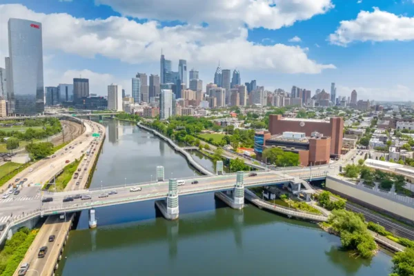 Philadelphia skyline and Schuylkill River with bridge traffic and Center City buildings under a partly cloudy sky.