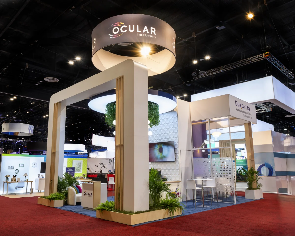 Angled view of the Ocular Therapeutix exhibit showing its large entrance arch, circular hanging sign, illuminated canopy, greenery, and meeting area