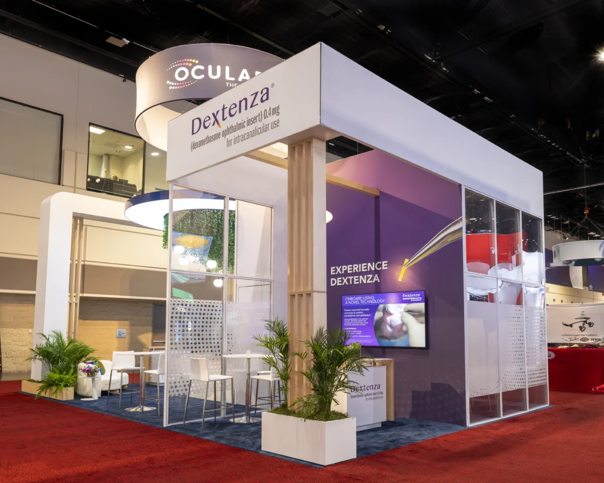 Side view of the Ocular Therapeutix exhibit showing a Dextenza display, glass-walled meeting area, plants, and circular hanging sign