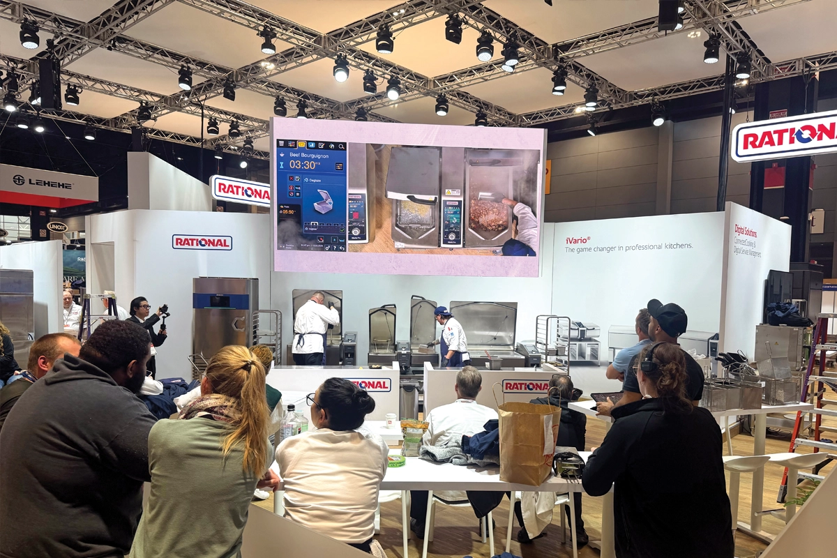 Attendees watch a RATIONAL USA cooking demonstration beneath a large LED display