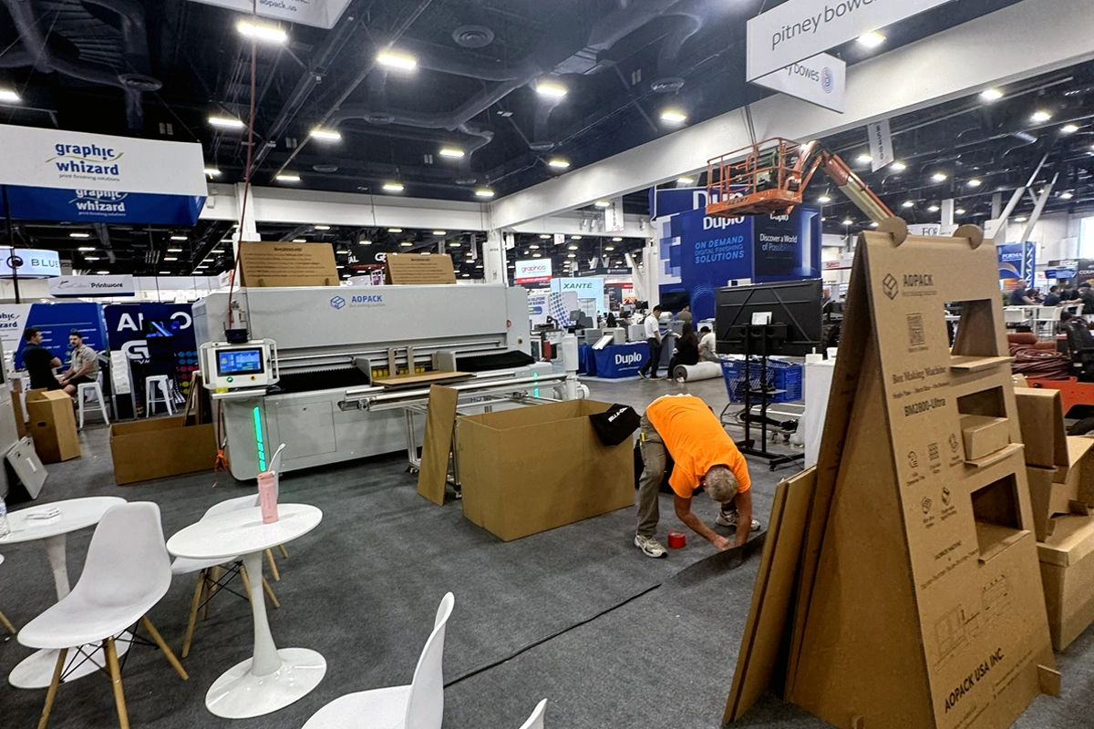 A worker secures flooring beside unpacked exhibit materials and equipment on a tradeshow floor.