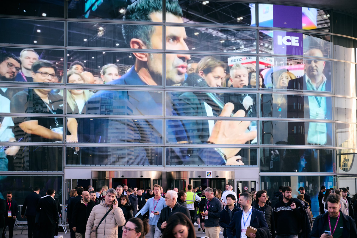 ICE attendees walk beneath a large video display showing a speaker holding a microphone on the exhibition floor.