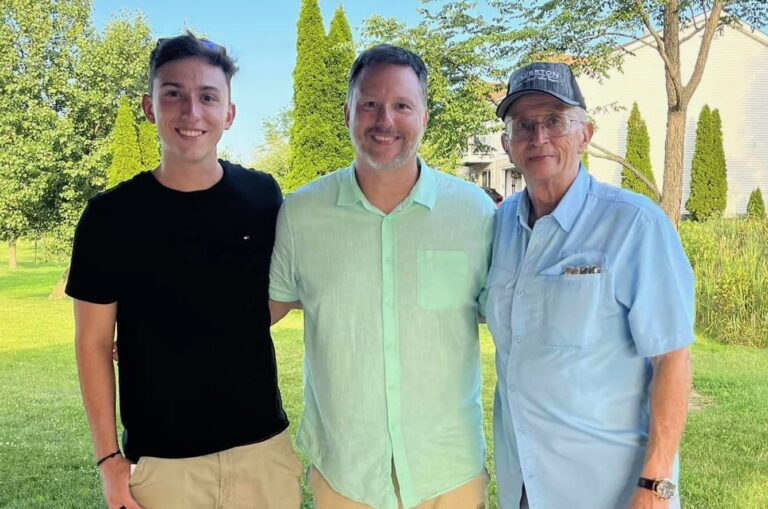 Mike Drobick, right, stands outdoors with two men, wearing a light blue shirt, glasses and a baseball cap.
