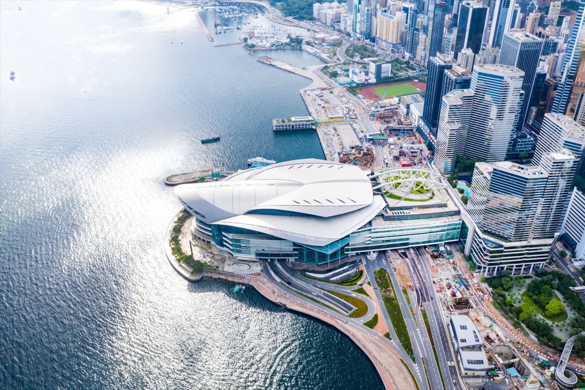 Aerial view of HKCEC’s curved roof beside Victoria Harbour and surrounding high-rise buildings.