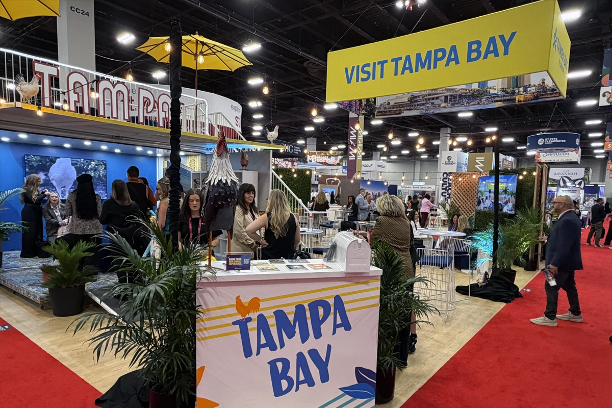 Attendees gather at Visit Tampa Bay’s exhibit beneath a yellow hanging sign, surrounded by plants, seating and an upper-level terrace.