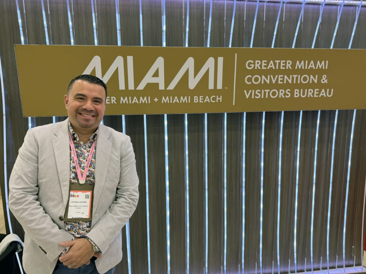 Christian Jaramillo smiles in a light gray jacket beside Greater Miami Convention & Visitors Bureau signage.