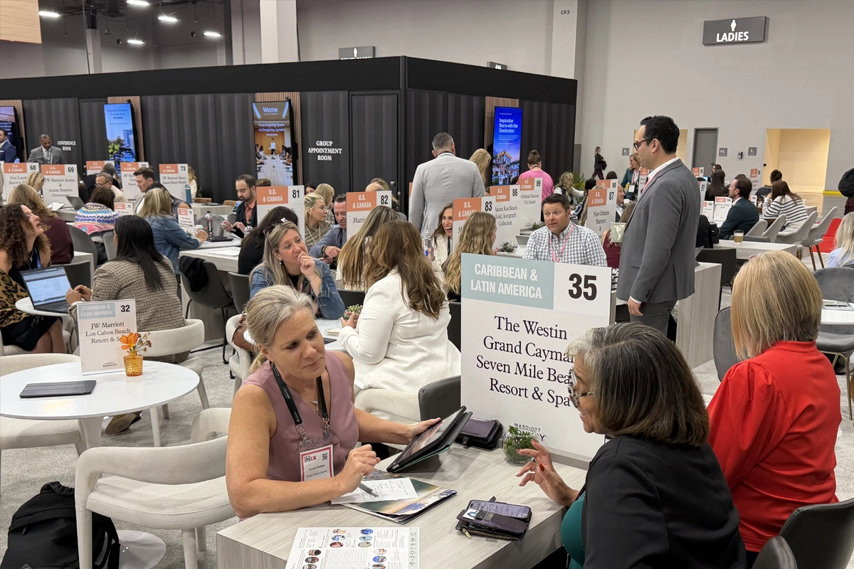 Attendees meet across tables marked with hotel names and regional signs on the IMEX America show floor.