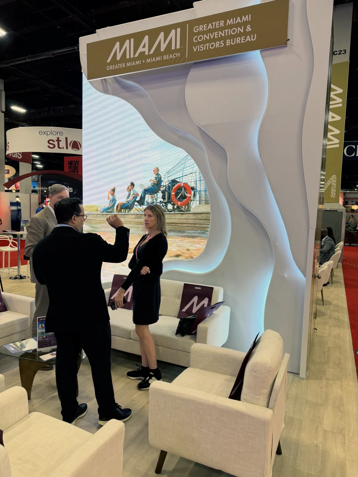 Attendees talk beside lounge seating and a large video display framed by curved, illuminated walls at Greater Miami’s exhibit.