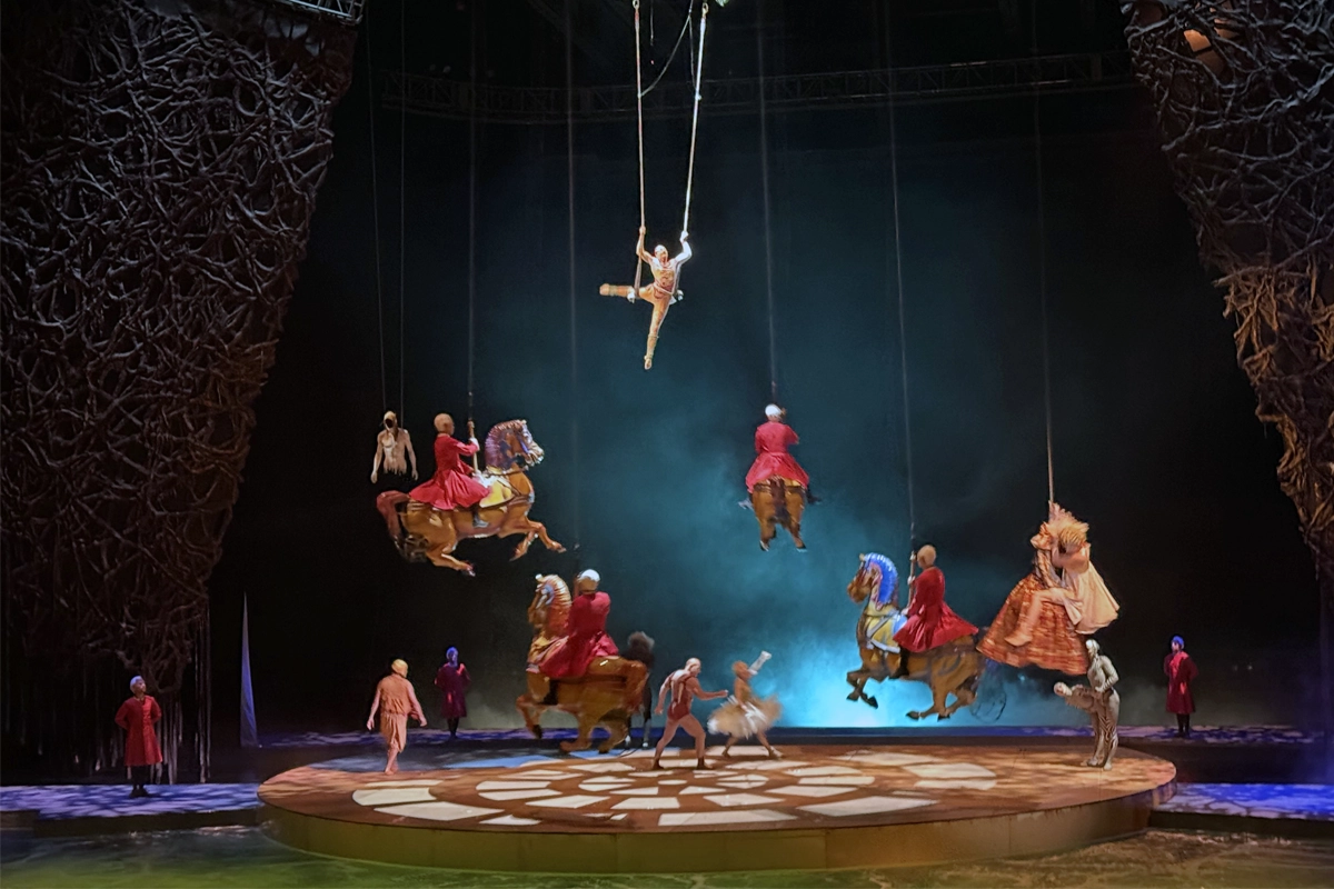 An aerial performer hangs above a circular stage as riders on suspended horse figures float over other performers.