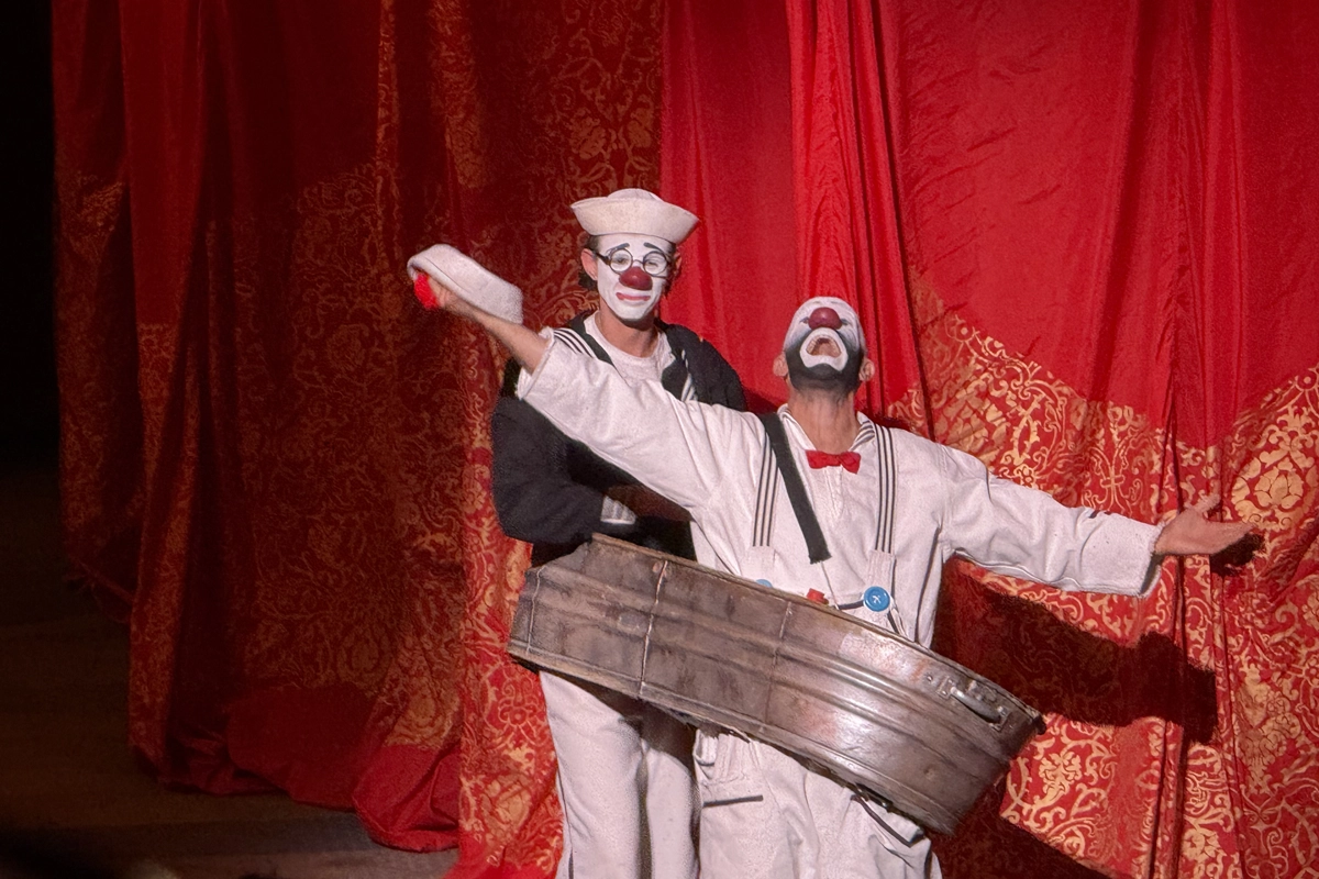 Two red-nosed sailor clowns pose before a red-and-gold curtain, one standing inside a metal tub with his arms outstretched.
