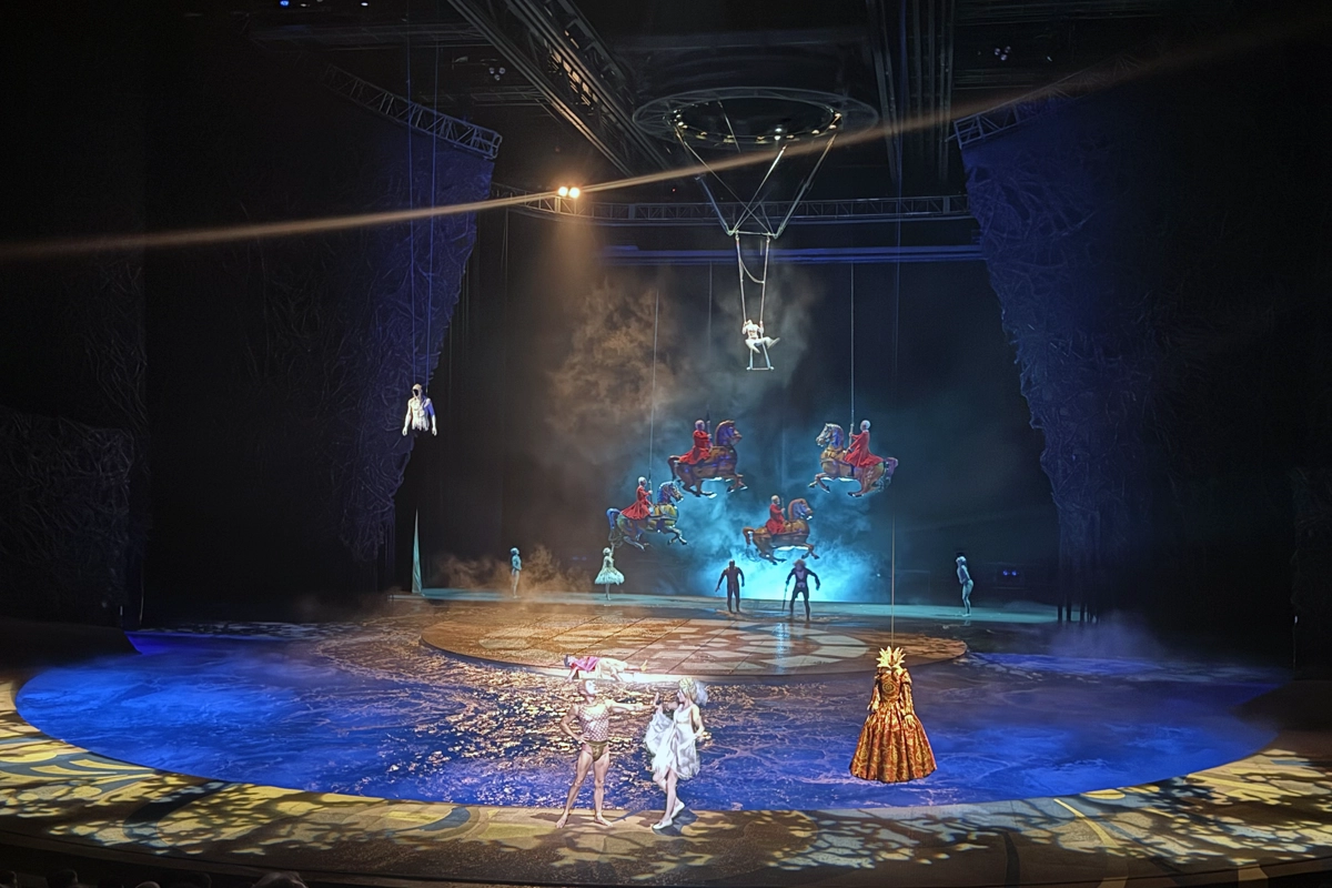 Wide view of Bellagio’s “O” stage with blue-lit water, aerial performers and suspended horse figures above a circular platform.