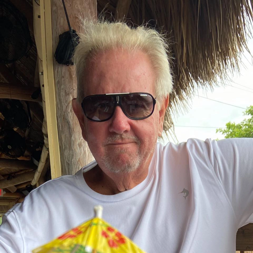 Tom Bacha wearing dark sunglasses and a white T-shirt while relaxing beneath a thatched roof.