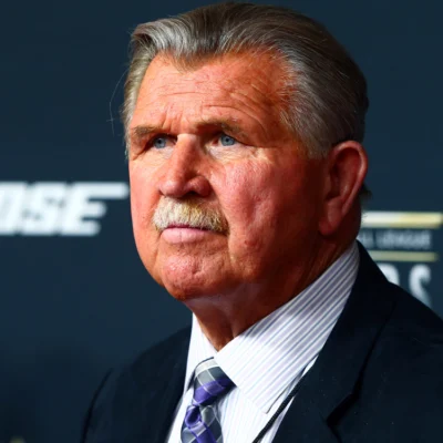 Mike Ditka wears a dark jacket, striped shirt and patterned tie against a dark backdrop.