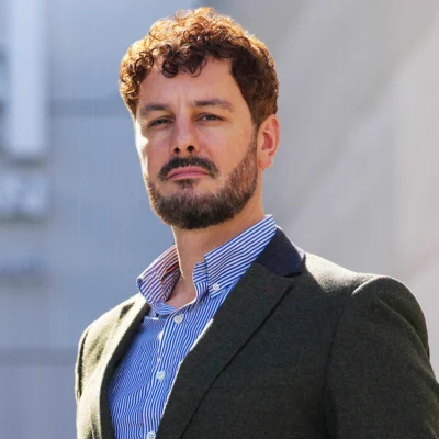 Sam Gibson poses in a dark jacket and blue striped shirt against a blurred background.