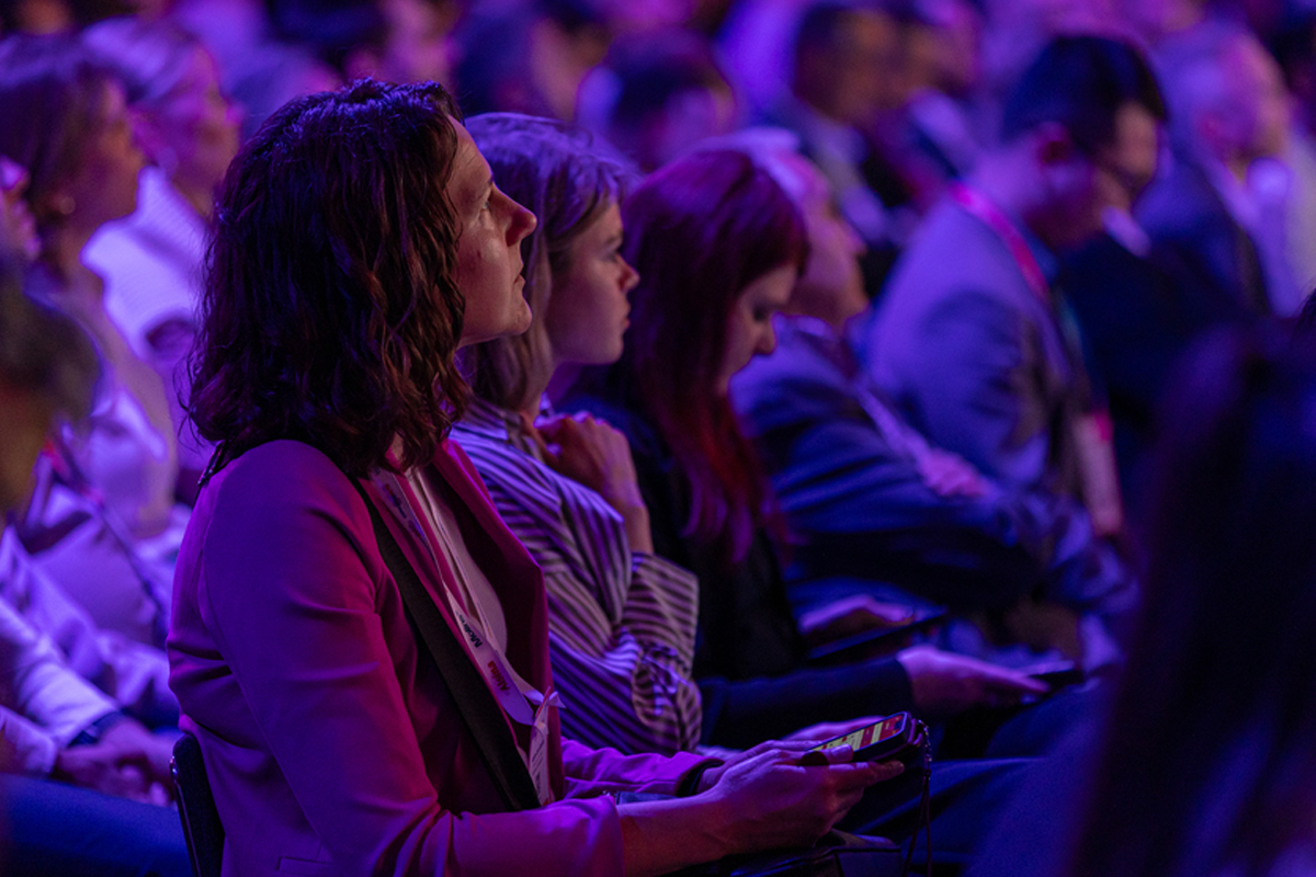 Conference attendees sit under purple lighting, with a woman holding a phone in the foreground.