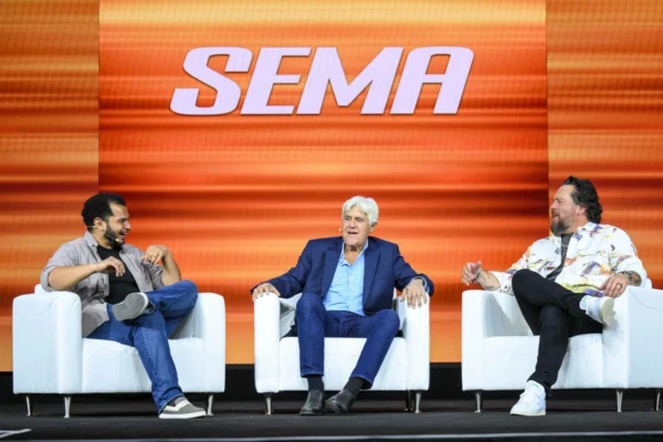 Jay Leno sits between two speakers in white armchairs beneath a large SEMA logo.