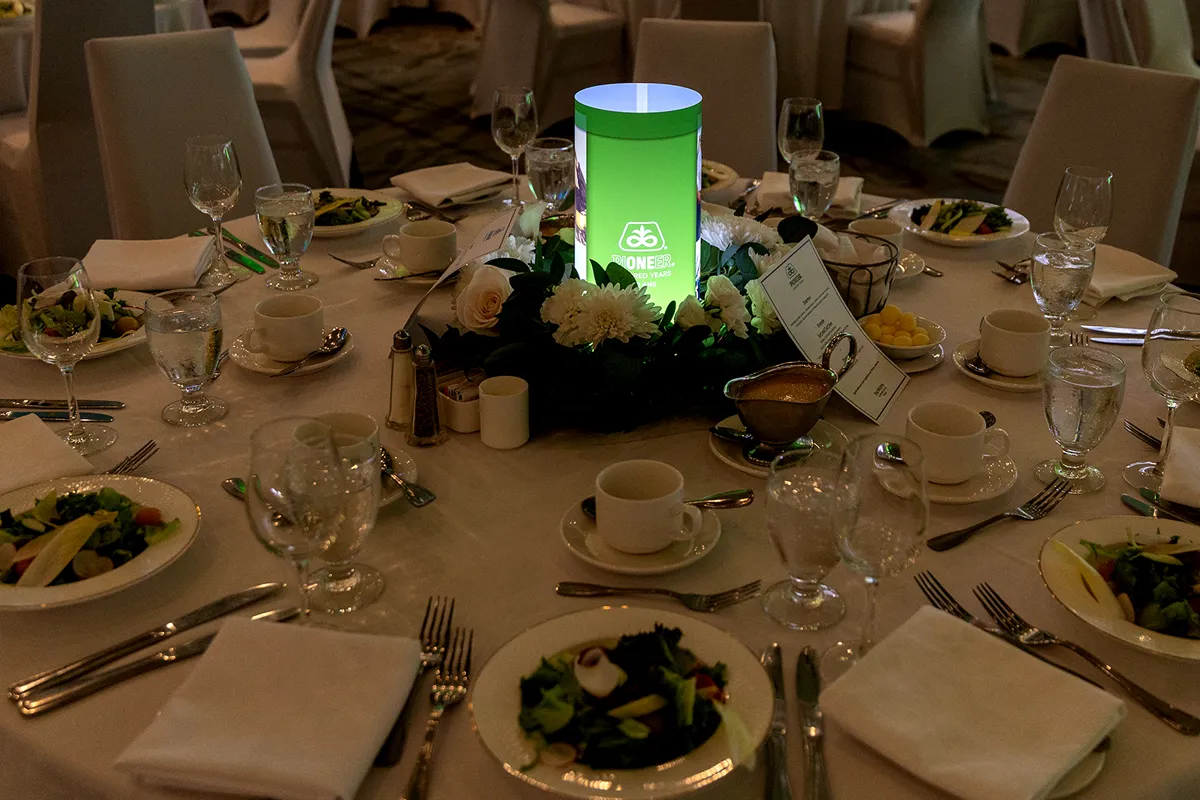 Green illuminated iLLUMITUBES centerpiece featuring Pioneer's 100th anniversary branding, surrounded by white flowers and banquet table settings.